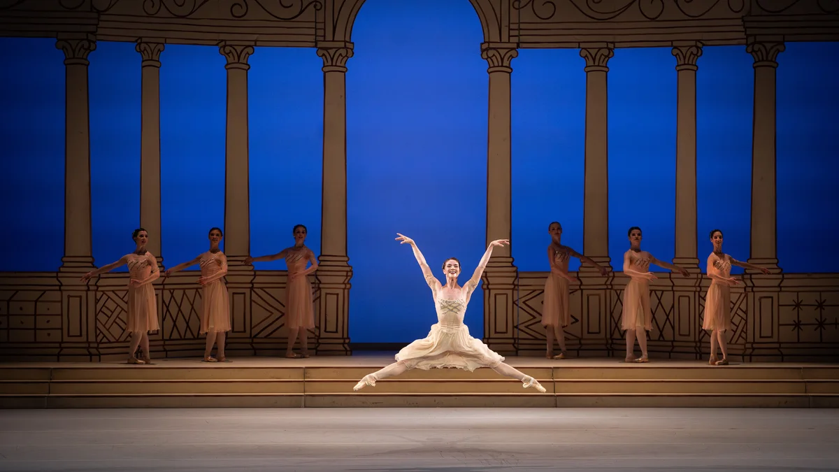 A ballerina leaps before the ensemble and blue-lit arches in Frederick Ashton’s Rhapsody.